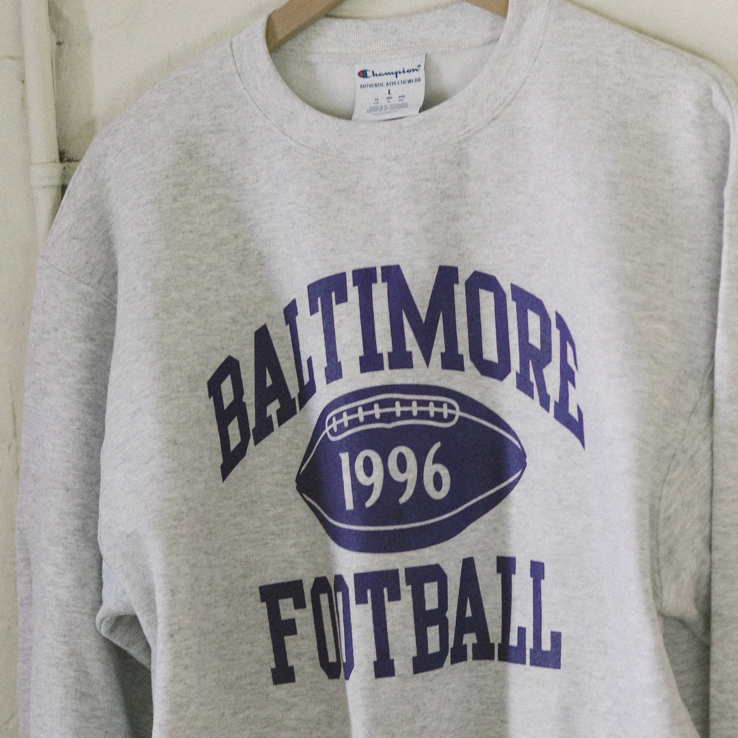 Light grey sweatshirt with purple Baltimore Football print, hanging on a wooden hanger in front of a white brick wall.